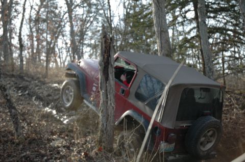 1998 Jeep TJ - A red 1998 Jeep TJ navigates a muddy trail surrounded by trees.