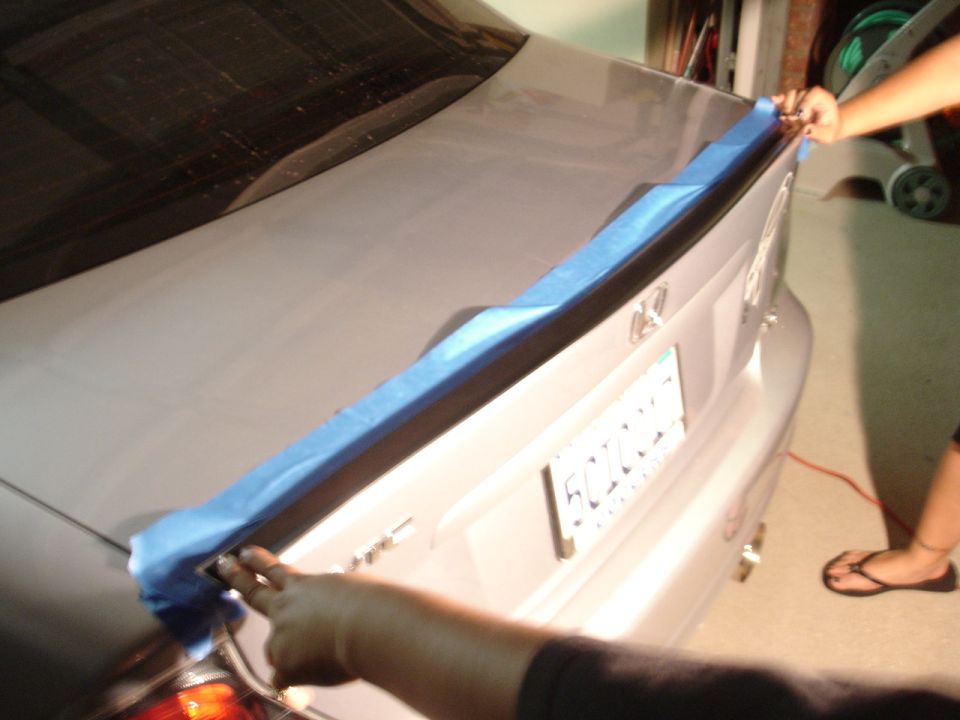 Hands applying blue tape to a car's trunk lid near the spoiler