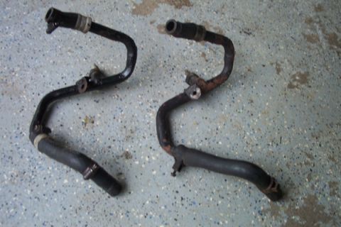 1995 Ford Taurus - Two engine coolant pipes, one black and one rusted, lie on a speckled floor.