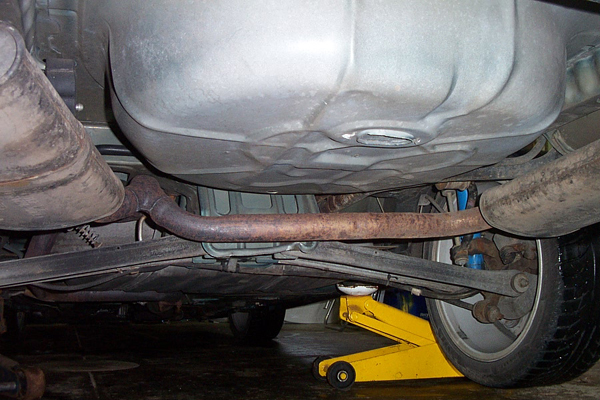 Underside view of a car's exhaust system, fuel tank, and suspension, with a yellow jack supporting the rear