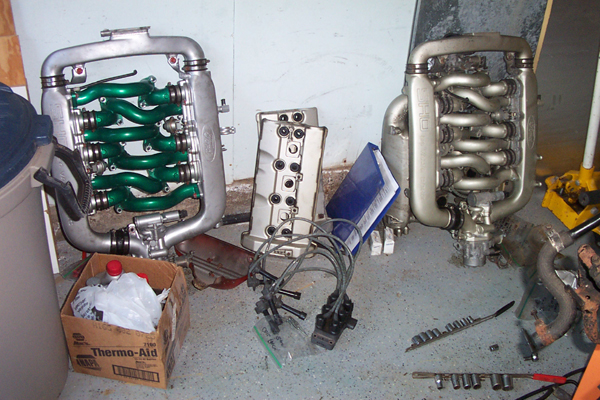 Two engine intake manifolds, one with green runners, sit on the floor next to engine parts and a box