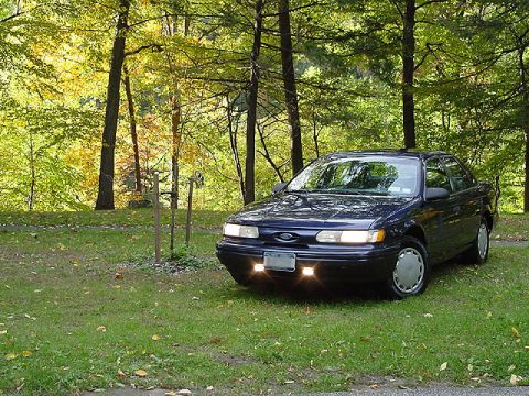 1993 Ford Taurus - A dark blue 1993 Ford Taurus sedan is parked on a grassy area surrounded by trees in the fall.