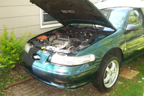 1995 Ford Taurus - The engine bay of a green sedan is exposed, with the hood propped open. The car appears to be a 1995 Ford Taurus SHO.