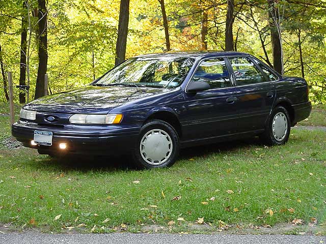 Dark blue Ford four-door sedan parked on grass in front of autumn trees, oval Ford badge on grille