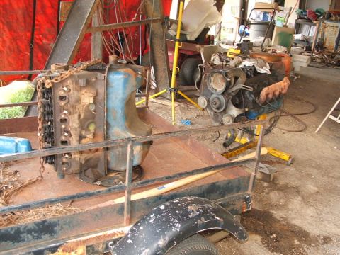 1966 Ford Ranchero - A disassembled V8 engine sits on an orange engine stand inside a shed, with a bare cylinder head and blue-painted intake manifold resting o…