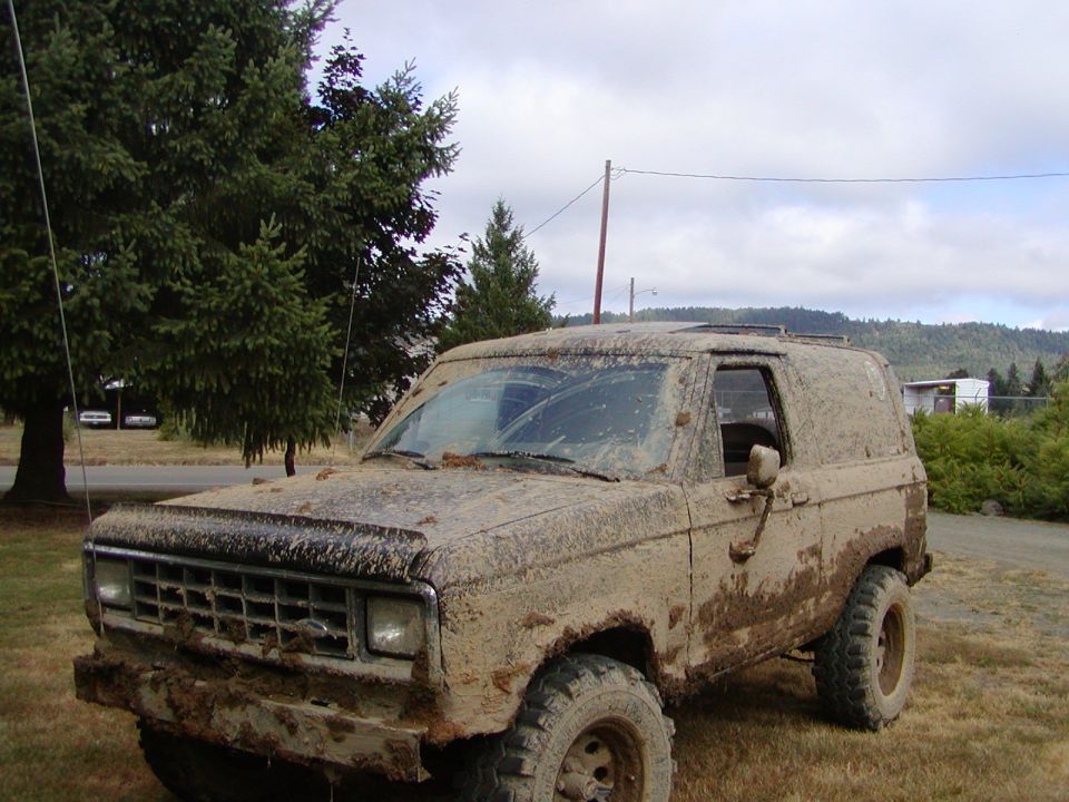 Ford SUV completely caked in dried mud, lifted on knobby tires, parked on grass under overcast sky