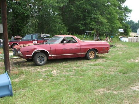 1972 Ford Ranchero - A red pickup truck coupe sits in a yard with a black pickup truck behind it, both appearing to be in disrepair.