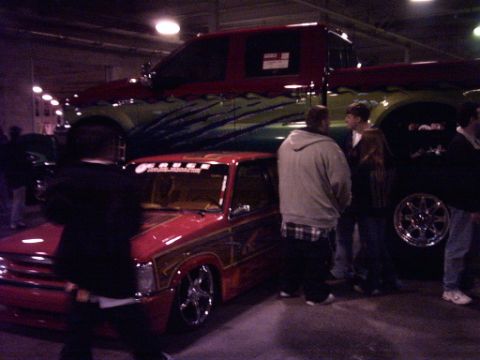 1995 Ford Probe - Two modified pickup trucks are displayed indoors with onlookers gathered around.