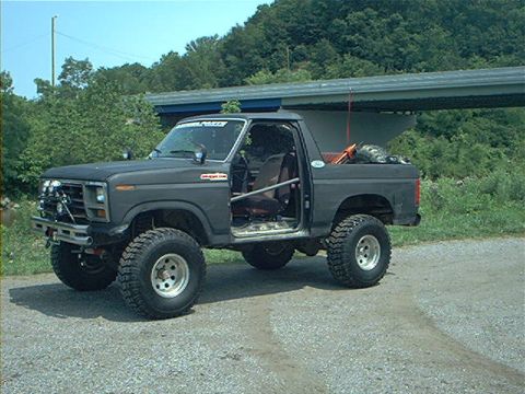 1986 Ford Bronco - This modified Ford Bronco sits parked on a gravel surface with trees and a covered structure in the background.