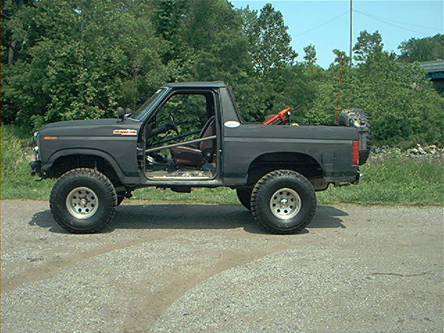 Dark gray Ford Bronco with doors removed, off-road tires, and roll cage, parked on a gravel road