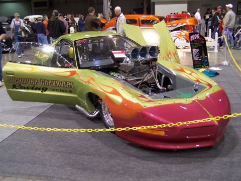 1995 Ford Probe - A custom drag car with a prominent engine and flame graphics is displayed indoors, with a sign indicating '1996'.