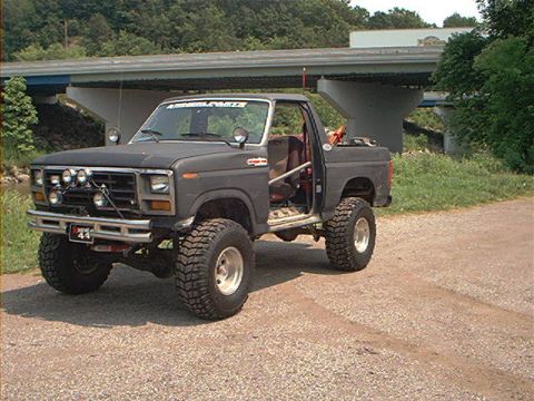 1986 Ford Bronco - Modified Ford Bronco with off-road tires and roll cage is parked on a gravel surface under a bridge.
