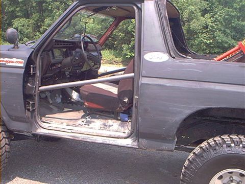 1986 Ford Bronco - The interior of a modified Ford Bronco is visible with the driver's door open, revealing a roll cage and seats.