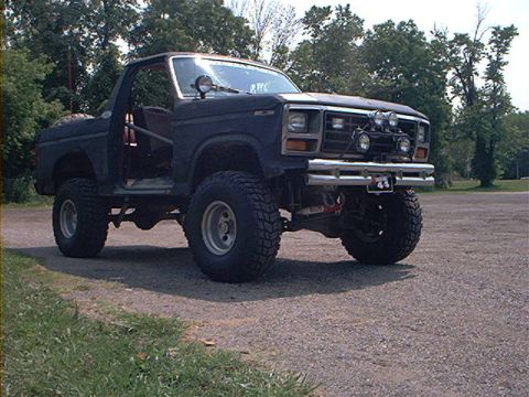 1986 Ford Bronco - This dark SUV, modified for off-road use, is parked on a gravel surface with trees in the background.