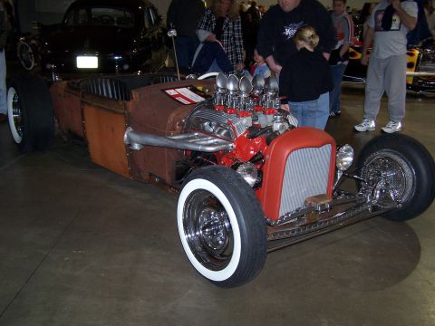 1995 Ford Probe - A heavily modified hot rod with a V8 engine and whitewall tires is displayed indoors, with other vehicles and people in the background.