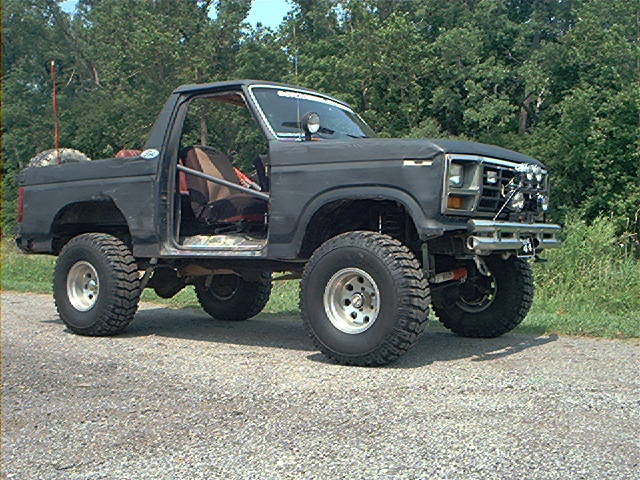 Modified 1980s Ford Bronco with roll cage and off-road tires parked on gravel