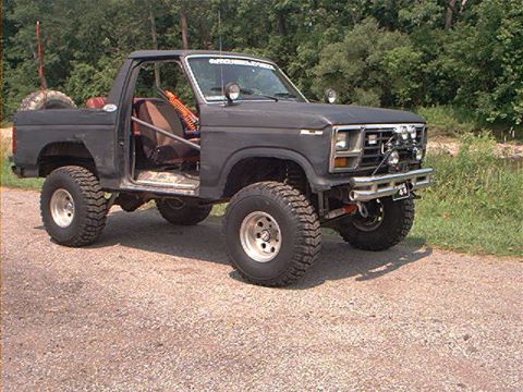 1986 Ford Bronco - This modified SUV is parked on a gravel road with trees and water in the background.