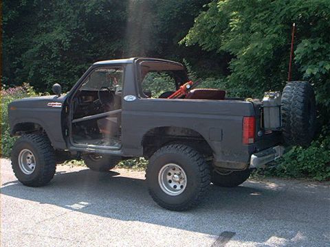1986 Ford Bronco - This modified Ford Bronco is parked on a paved surface with trees in the background.