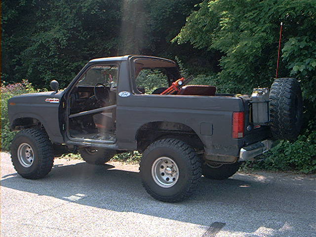 Dark gray Ford Bronco with roll cage, off-road tires, and spare tire on the back. Doors are removed