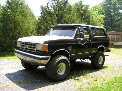 1988 Ford Bronco - This lifted 4x4 Bronco sits on a gravel driveway with trees and a house in the background.