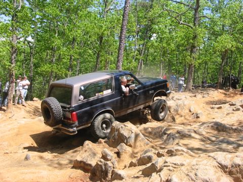 1988 Ford Bronco - A lifted black Ford Bronco navigates a rocky off-road course with spectators watching.