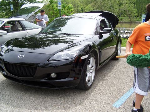 1995 Ford Probe - A black Mazda RX-8 is parked next to another car with its hood open, with a person holding a car duster nearby.