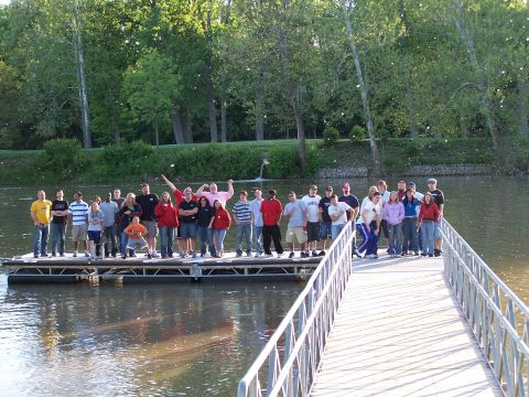 1995 Ford Probe - A large group of people gathered on a floating dock near a tree-lined shore.