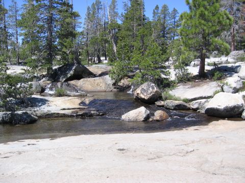 1994 Ford Bronco - A clear mountain stream winds through a rocky landscape dotted with pine trees under a bright blue sky.