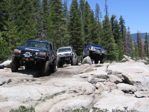 1994 Ford Bronco - Three off-road vehicles tackle a challenging rocky path surrounded by pine trees under a clear blue sky.