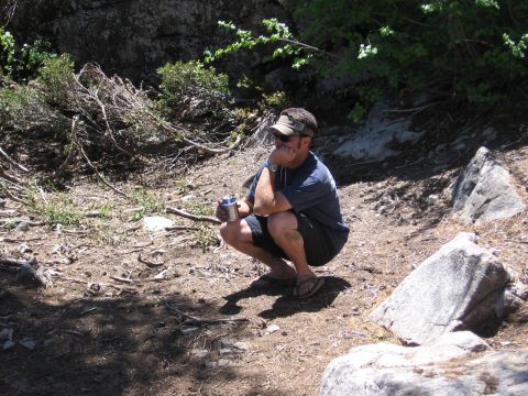 1994 Ford Bronco - A man takes a break while squatting in a rocky, wooded outdoor area, holding a beverage can.