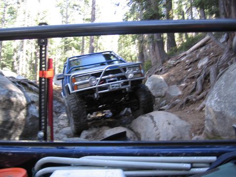 1994 Ford Bronco - Blue off-road SUV navigates a challenging rocky trail in a wooded area.