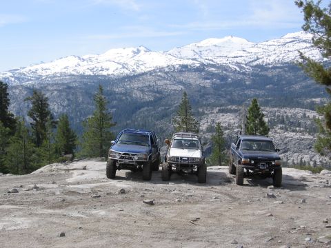 1994 Ford Bronco - Three off-road vehicles are parked on a rocky outcrop, with a vast, snow-covered mountain range visible in the distance under a clear blue…