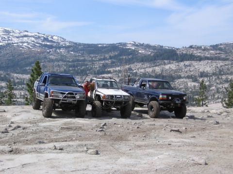 1994 Ford Bronco - Three modified trucks, including a 1994 Ford Bronco, are parked on a rocky, mountainous terrain.