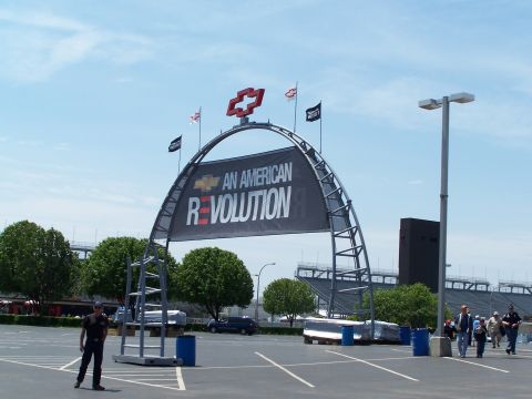 1995 Ford Probe - An archway with a Chevrolet logo and banner reading 'AN AMERICAN REVOLUTION' stands over a parking lot with a stadium in the background.
