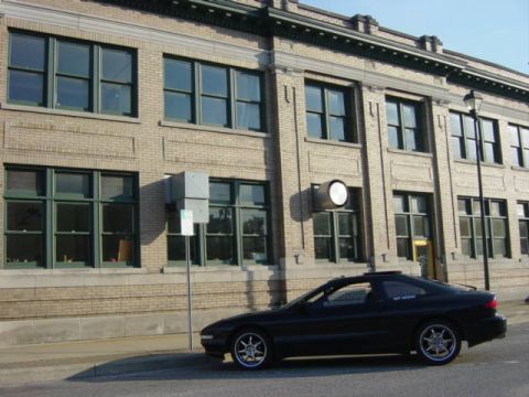 1995 Ford Probe - A black coupe is parked on the street in front of a brick building with many windows.