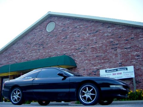 1995 Ford Probe - A black 1995 Ford Probe is parked in front of a brick building with shelter hours posted on a sign.
