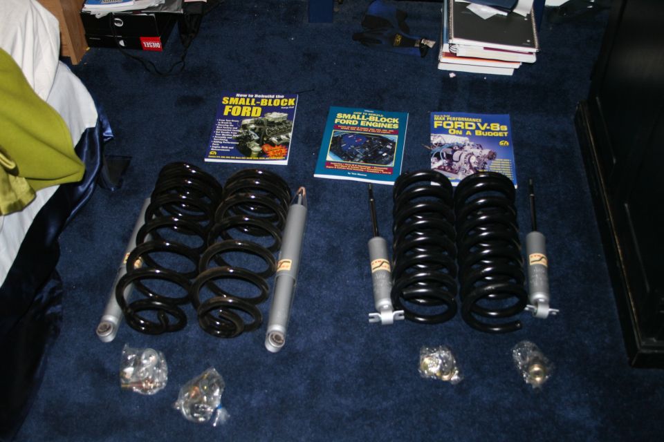 Four coil springs and four shock absorbers laid out on a dark blue carpet next to automotive repair books