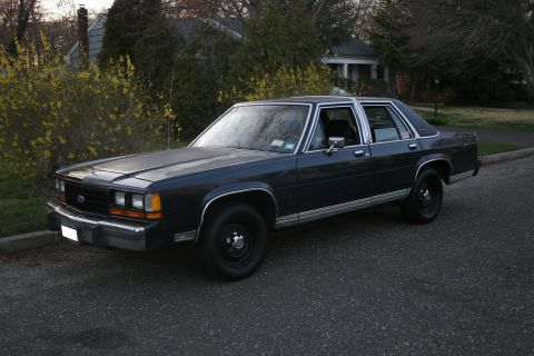 1989 Ford Crown Victoria LTD - A dark blue sedan is parked on a street with houses and trees in the background.