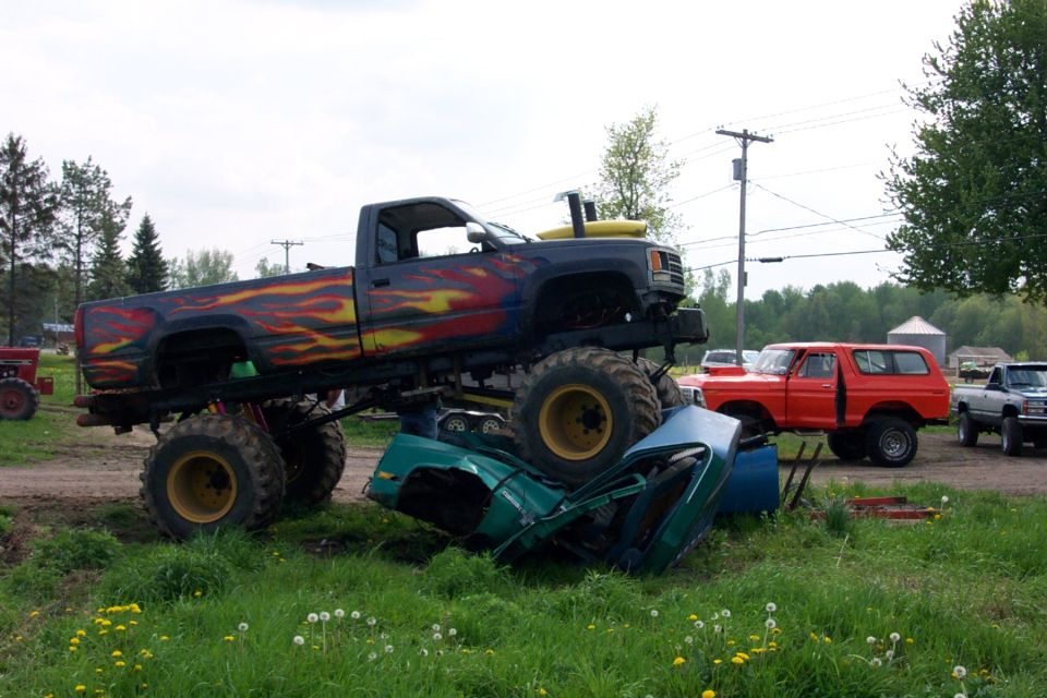Monster truck with flame decals crushing a green car, with a red SUV and a pickup truck in the background