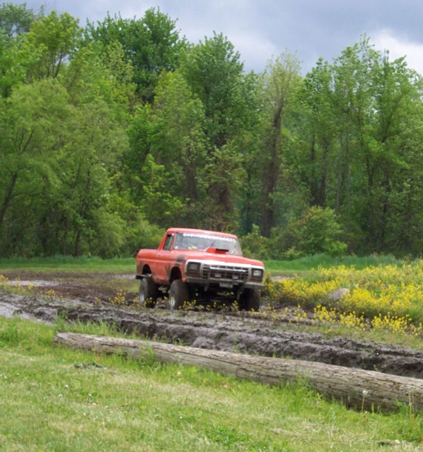 Red 1979 Ford F-Series pickup truck driving through mud with yellow flowers and trees in background
