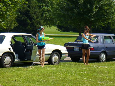 1991 Ford Probe - Two women are preparing for a water gun fight next to two cars on a sunny day.
