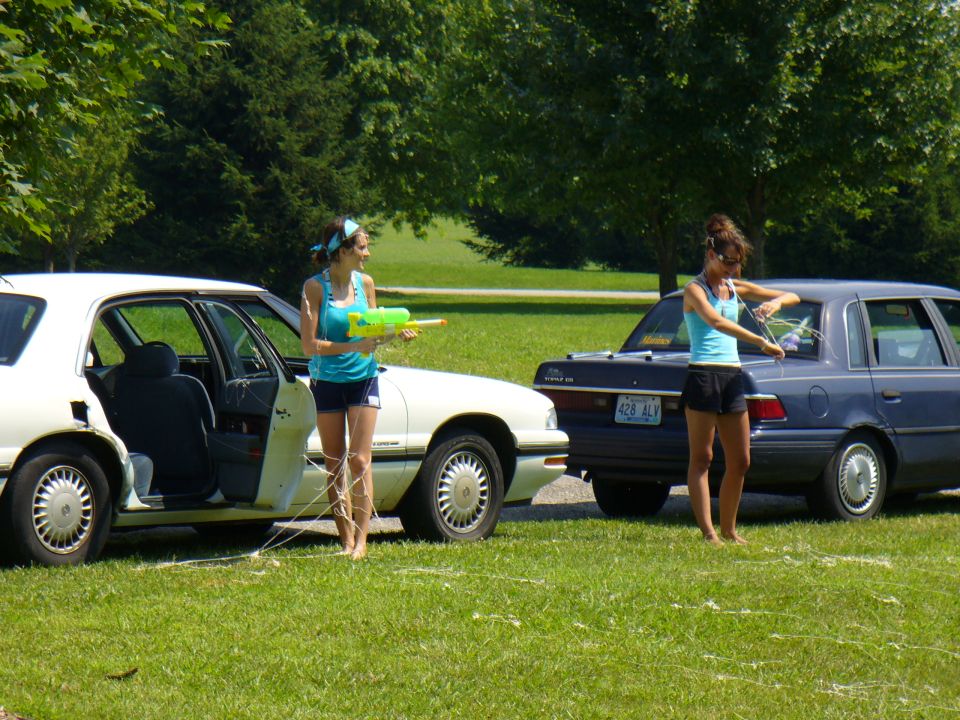 Two women stand by cars in a grassy area, one holding a water gun
