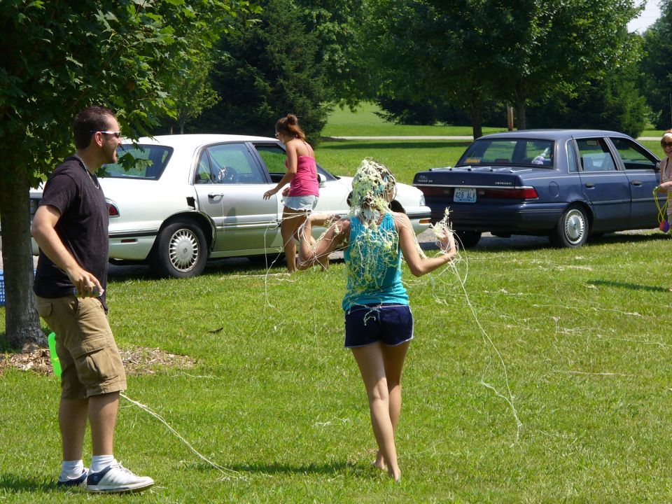People at an outdoor gathering with two cars in the background. One person has something green on their head