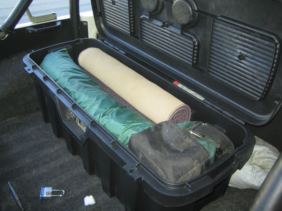 Open black storage box in vehicle interior containing green sleeping bag and rolled tan mat