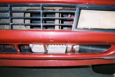 1991 Ford Probe - Close-up of a dark red car's front bumper and lower grille opening, where a silver heat-exchanger core and a black ribbed intake hose with…