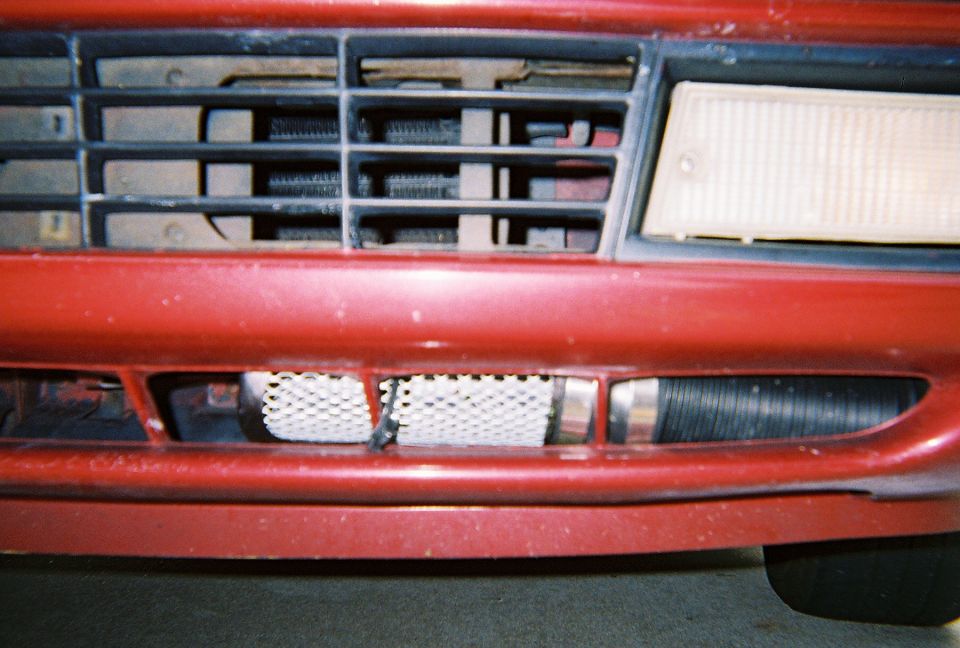 Close-up of red car's lower front bumper showing heat-exchanger core and ribbed intake hose behind grille slats
