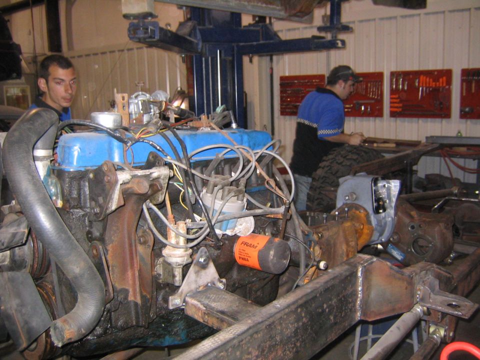 Engine and chassis assembly in a workshop with two mechanics present. Fram oil filter visible