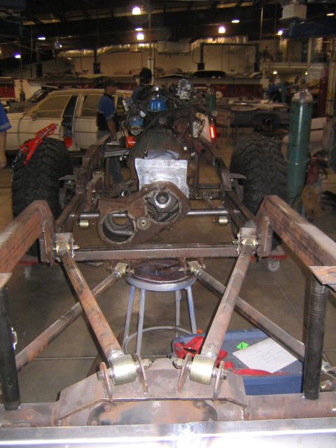 1982 Ford Bronco - This image shows the chassis of a vehicle, focusing on the engine and drivetrain components, with a partial view of a car in the background.