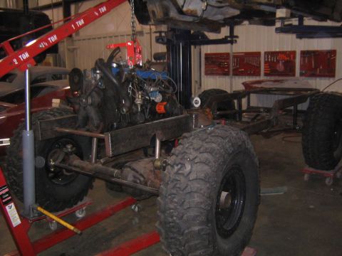 1982 Ford Bronco - Vehicle chassis and engine assembly suspended by a red engine hoist in a workshop setting.