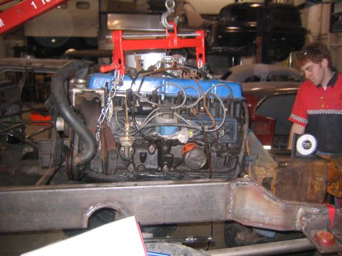 1982 Ford Bronco - Engine being lowered into a vehicle frame with a red engine hoist in a workshop setting.
